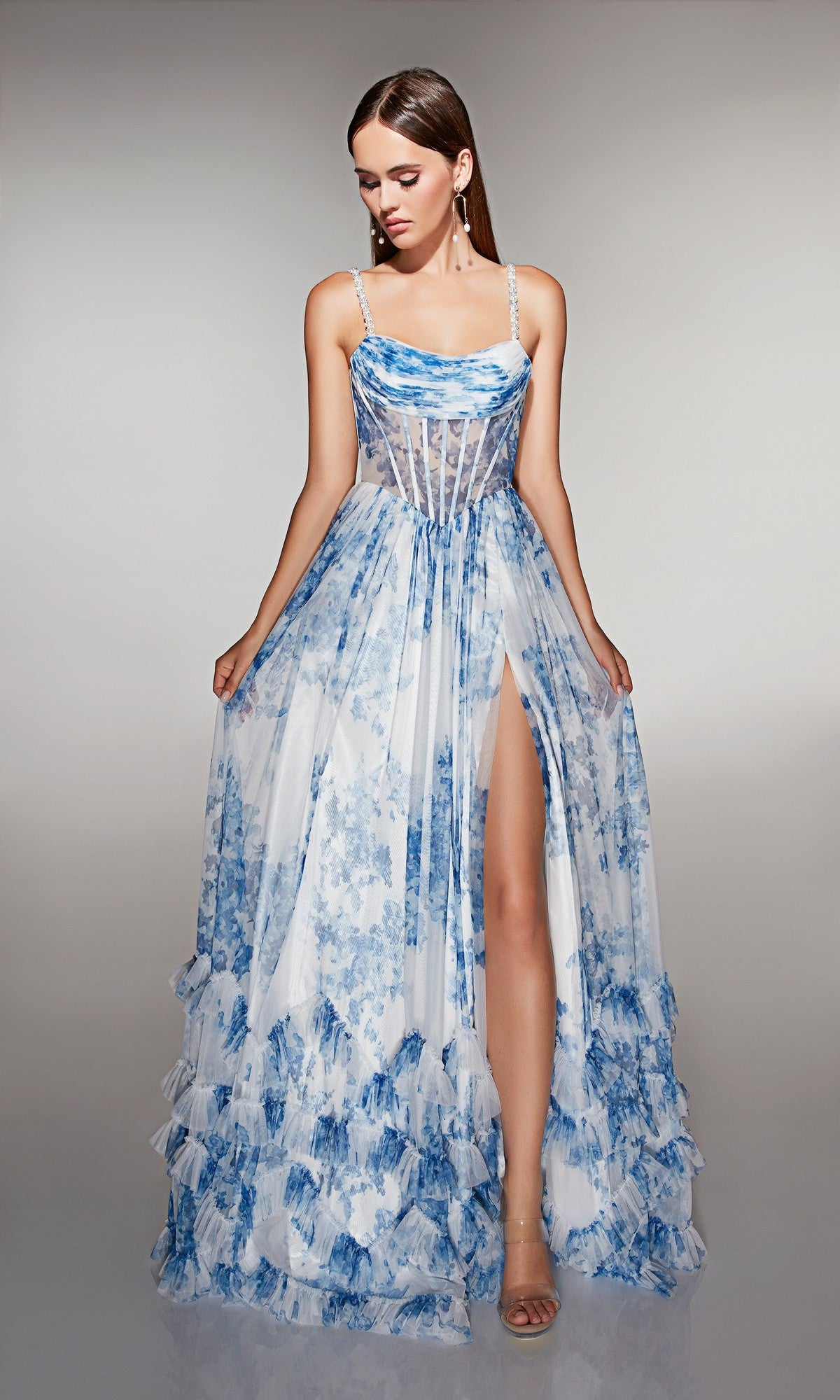 A woman wears the Alyce 62202 long prom dress, featuring a blue and white floral print, high slit, and sheer corset bodice, standing against a plain background.